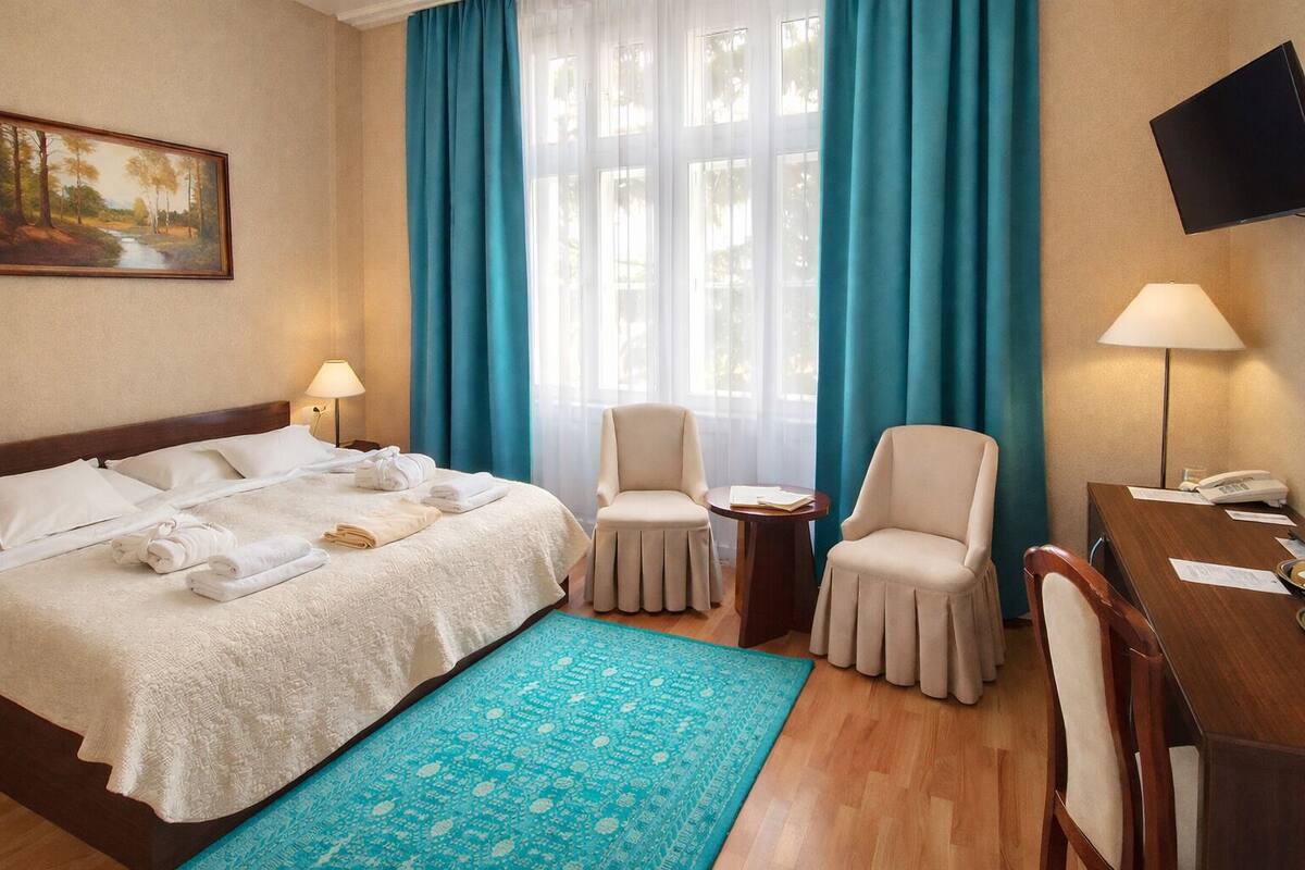 A hotel room with a bed, two chairs, a desk, a TV, and a painting on the wall.