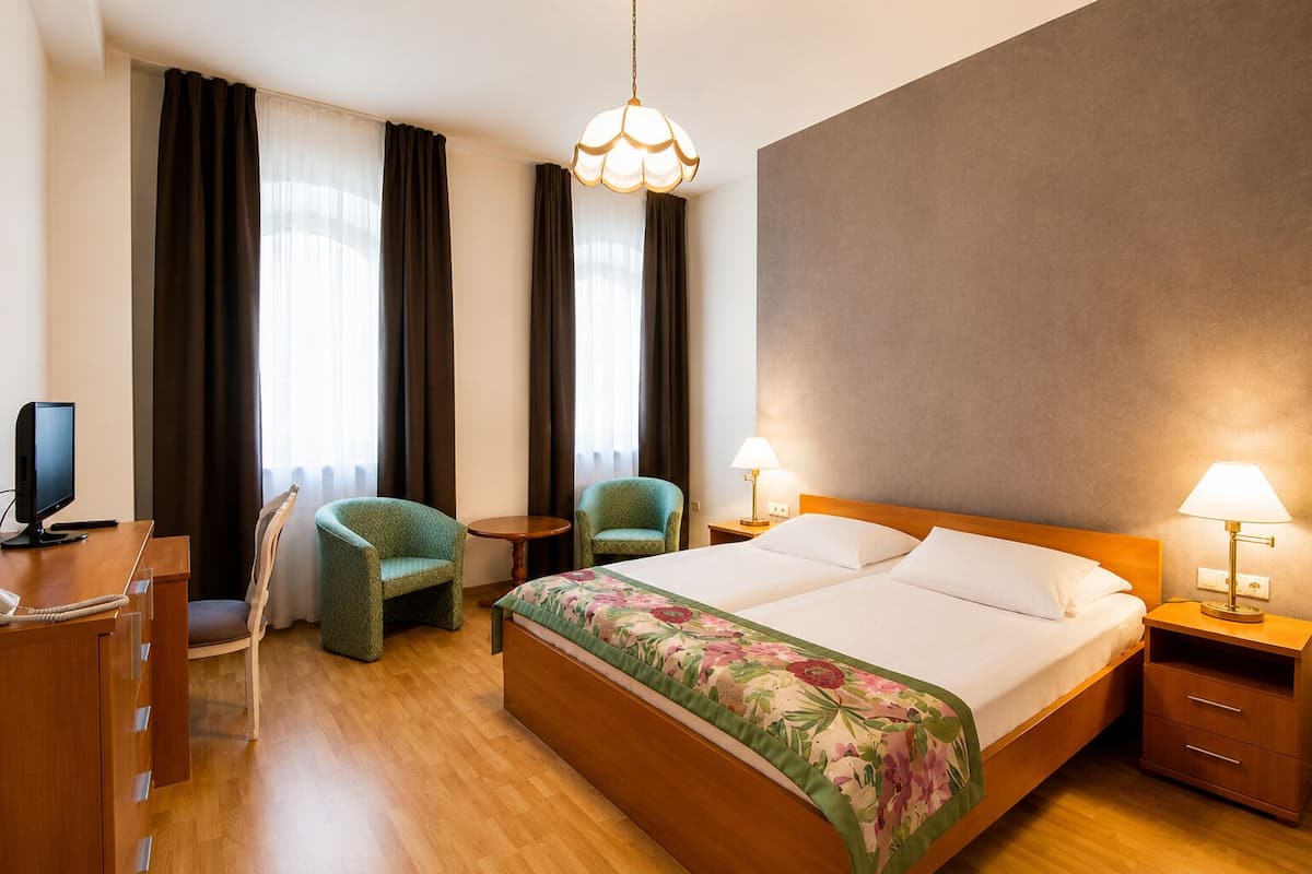A hotel room with a bed, two chairs, a desk with a computer, and a TV.