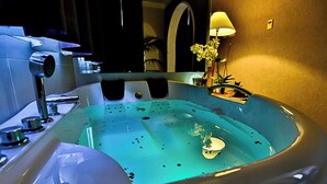 A large, modern bathtub with a built-in seat, a handheld showerhead, and a small table with a lamp. - Batthyány Kastélyszálló (Zalacsany)