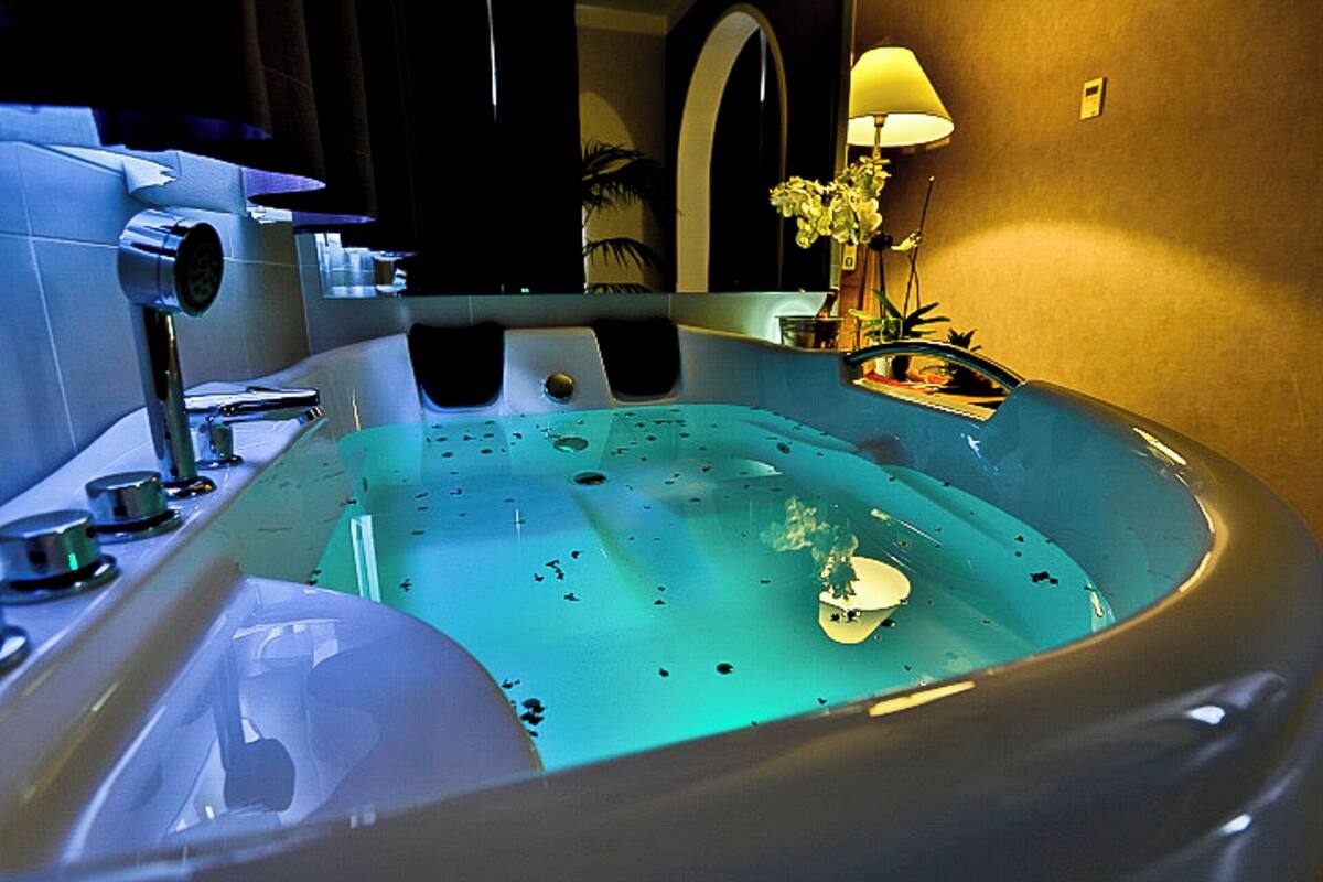 A large, modern bathtub with a built-in seat, a handheld showerhead, and a small table with a lamp.