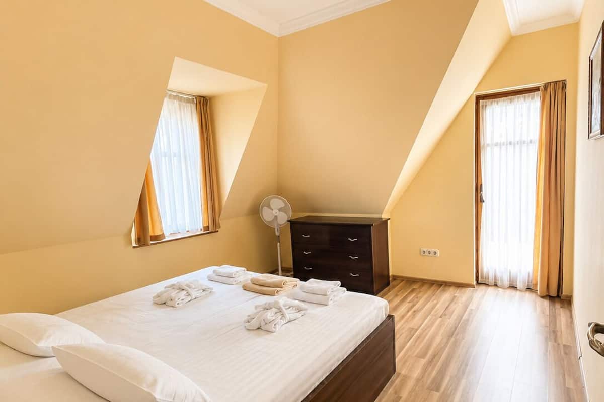 A bedroom with a large bed, a dresser, a fan, and a window with curtains.