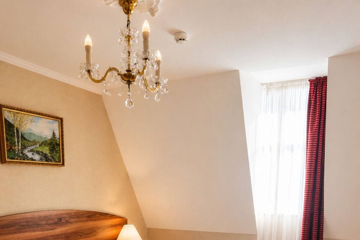 A bedroom with a wooden headboard, a patterned bedspread, a chandelier, a painting on the wall, and a window with curtains.
