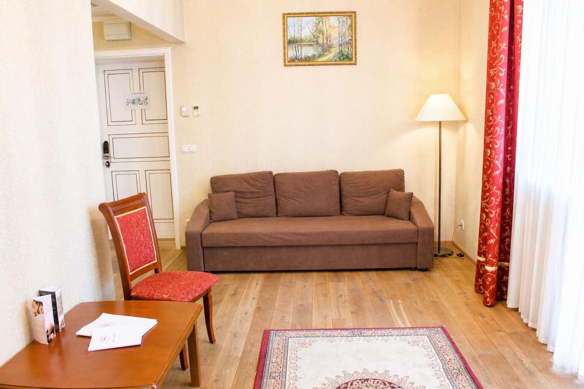 A living room with a brown sofa, a wooden chair with a red cushion, a wooden table with papers, a floor lamp, and a patterned rug.