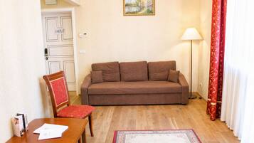 A living room with a brown sofa, a wooden chair with a red cushion, a wooden table with papers, a floor lamp, and a patterned rug.