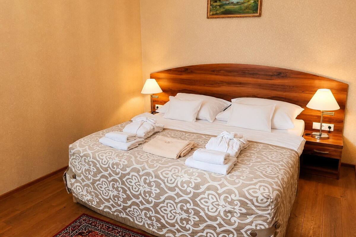 A hotel room with a wooden headboard, two bedside lamps, a bed with a patterned bedspread, and a painting on the wall.