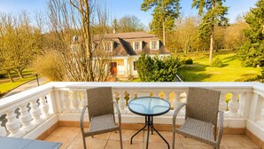 A balcony with wicker chairs and a glass table, overlooking a house and a well-maintained garden. - Batthyány Kastélyszálló (Zalacsany)