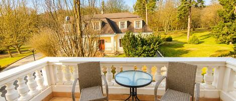 A balcony with wicker chairs and a glass table, overlooking a house and a well-maintained garden.