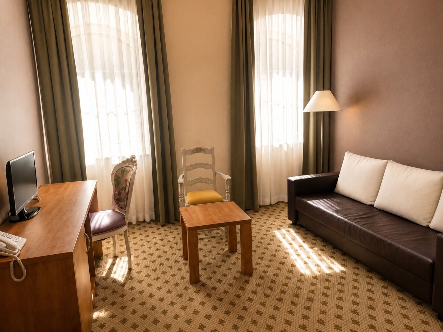 A hotel room with a wooden desk, a chair with a yellow cushion, a brown leather sofa, and a lamp.