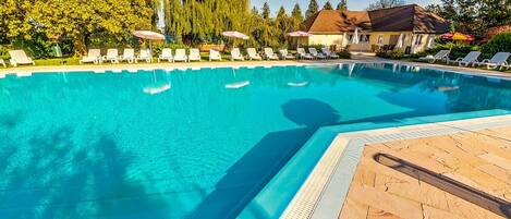 Una piscina cristalina con sillas de descanso y sombrillas, rodeada de árboles y un edificio.