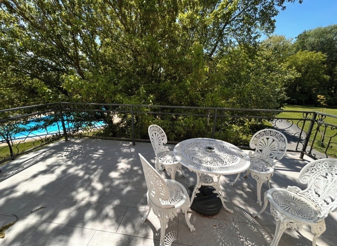 A patio with white metal chairs and a table, surrounded by trees and a pool.