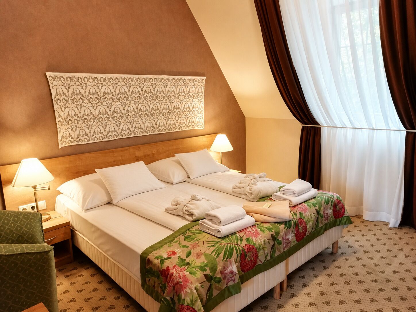 Une chambre d’hôtel avec un lit, deux lampes de chevet, une couvre-lit à motifs floraux, une tapisserie décorative et une fenêtre avec des rideaux.