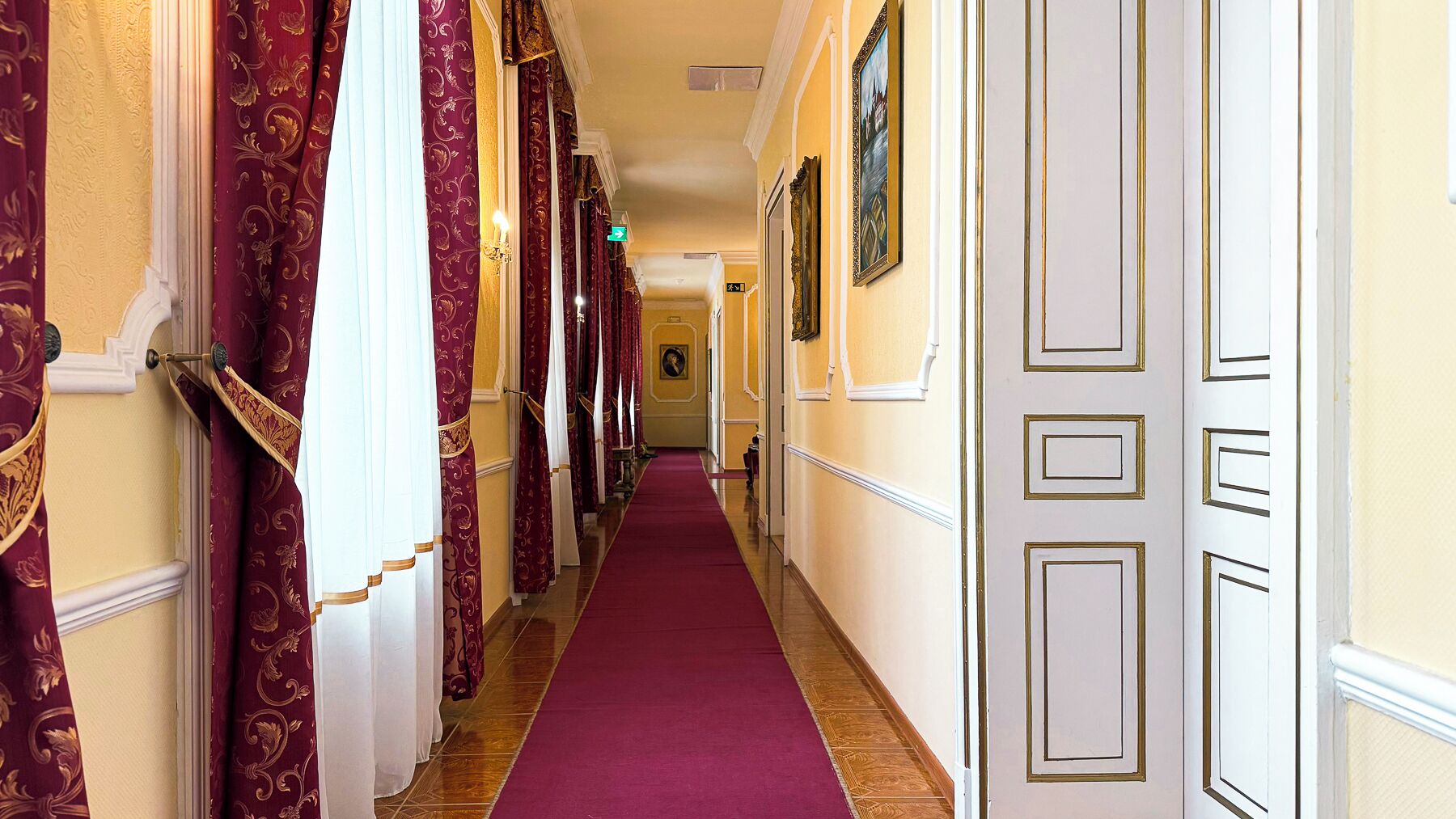 A hallway with a red carpet, patterned curtains, and a framed painting on the wall.