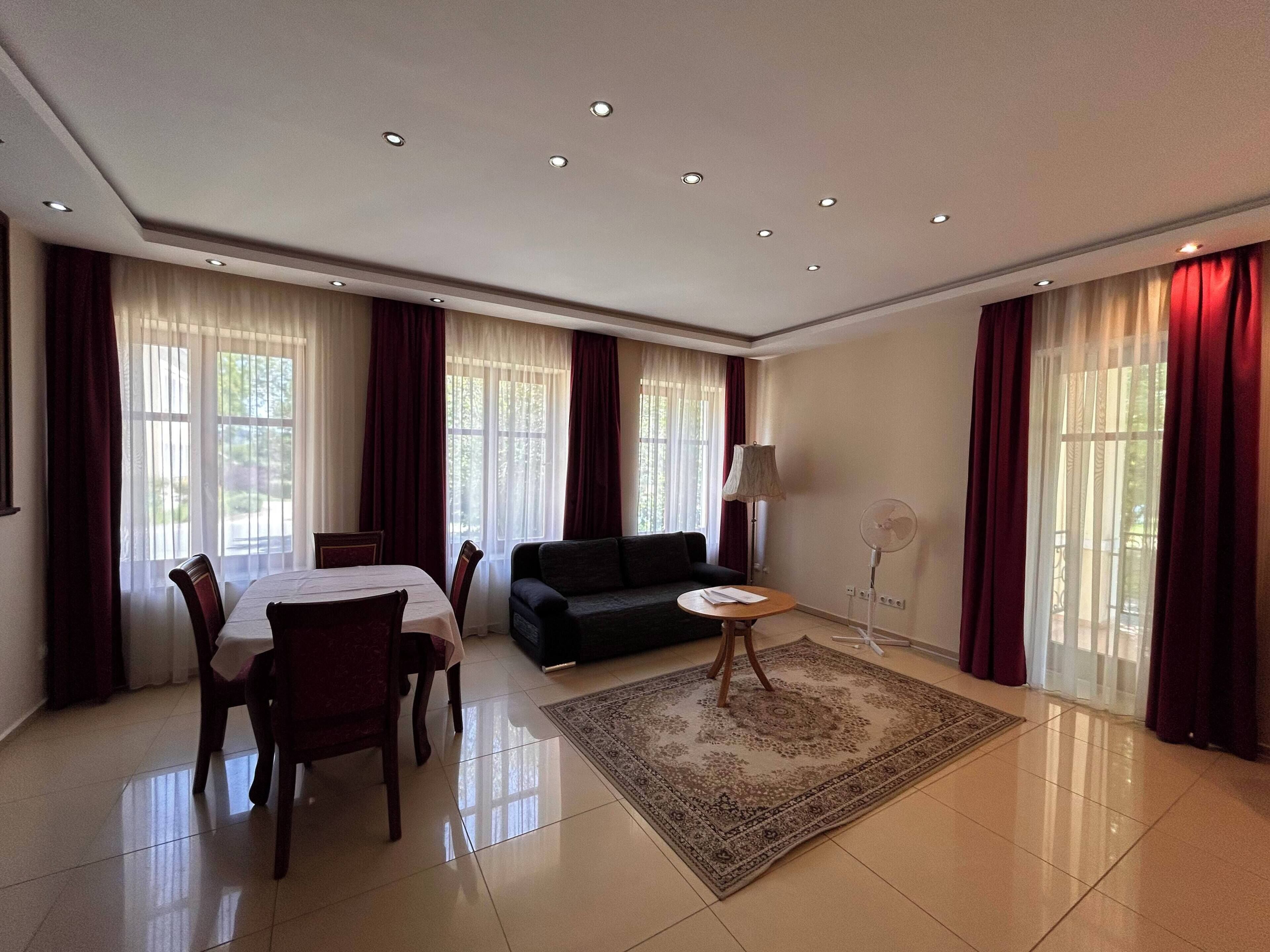 A spacious living room with a dining area, large windows with curtains, a sofa, a small table, and a standing fan.