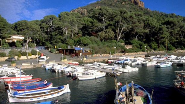 Un puerto deportivo con varias embarcaciones amarradas, una colina con un edificio en la cima, y una carretera que conduce al puerto.