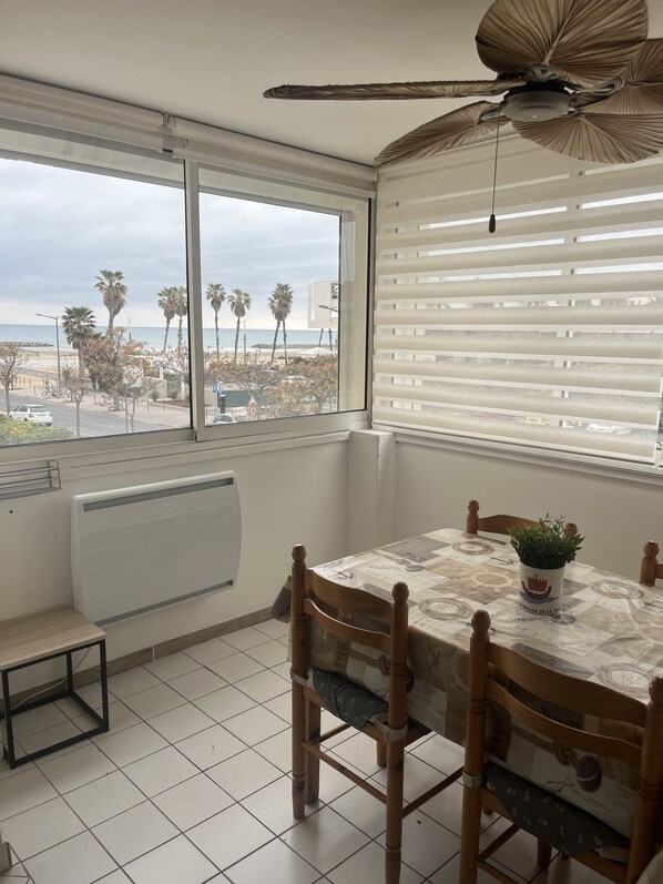 A dining area with a table, chairs, a ceiling fan, a window with blinds, and a view of the beach.