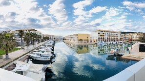 A marina with boats docked along a canal, surrounded by buildings with balconies and palm trees.
