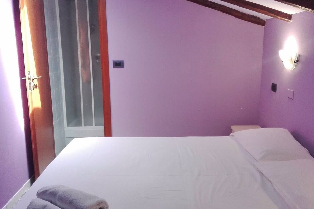 A room with a bed, a window with blinds, and a wall-mounted light fixture.