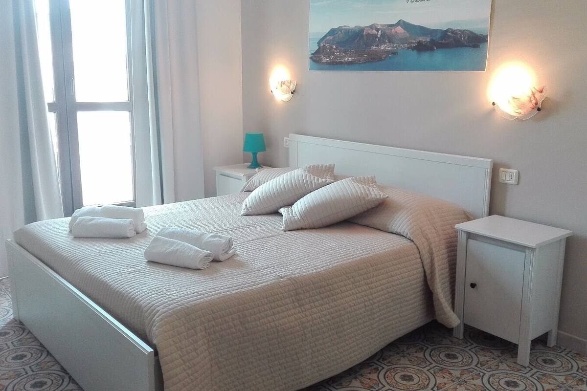 A neatly made bed with white linens and pillows, a bedside table with a lamp, and a wall-mounted light fixture.