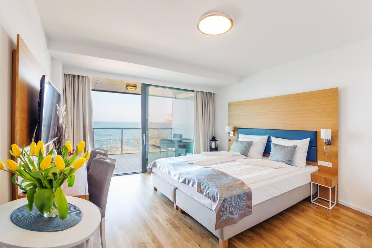 A modern hotel room with a large bed, a balcony with a view of the sea, a small table with a vase of flowers, and a TV mounted on the wall.