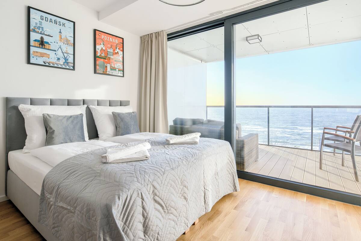 A bedroom with a large bed, a balcony with a view of the sea, and two posters on the wall.