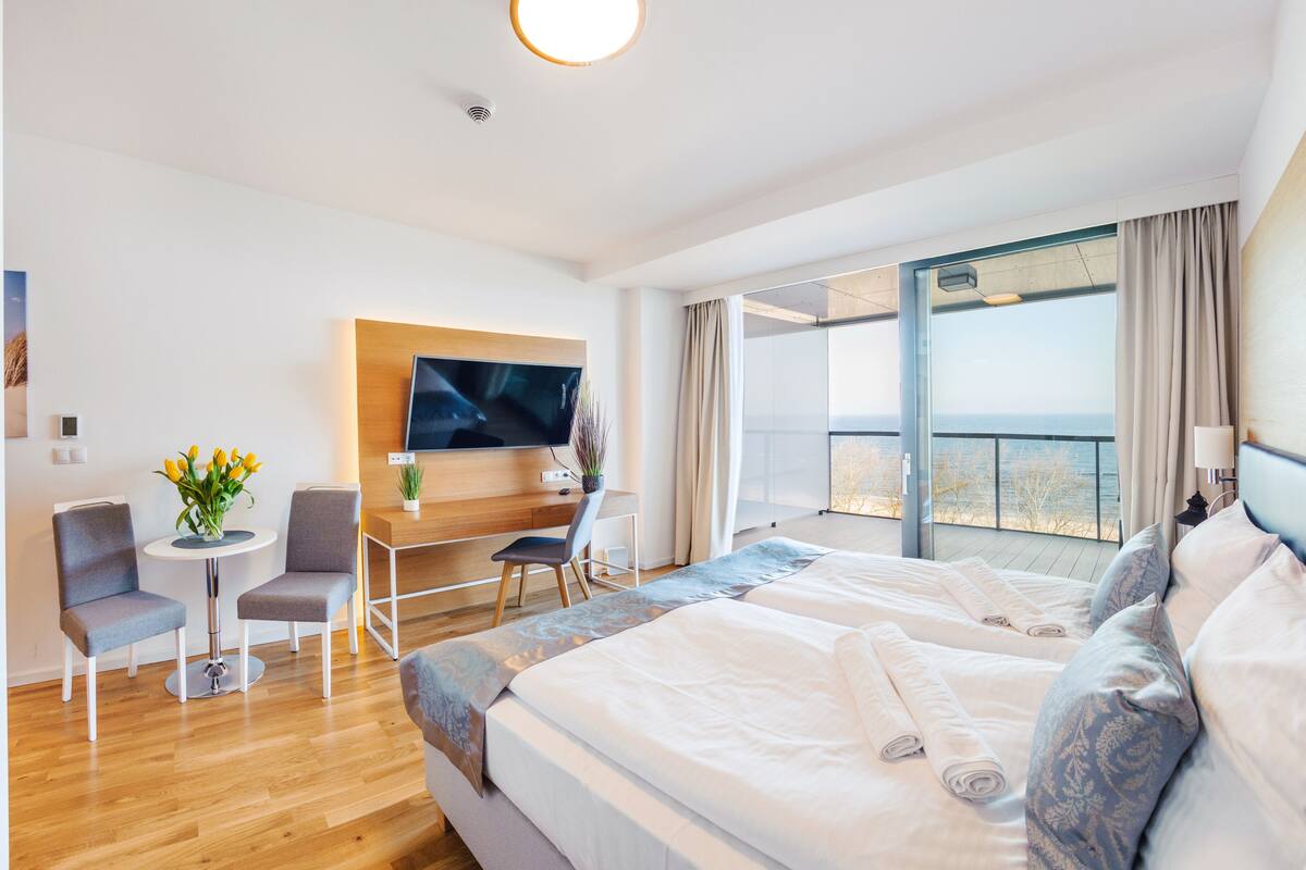 A modern hotel room with a large bed, a desk with a chair, a TV, and a balcony with a view of the sea.