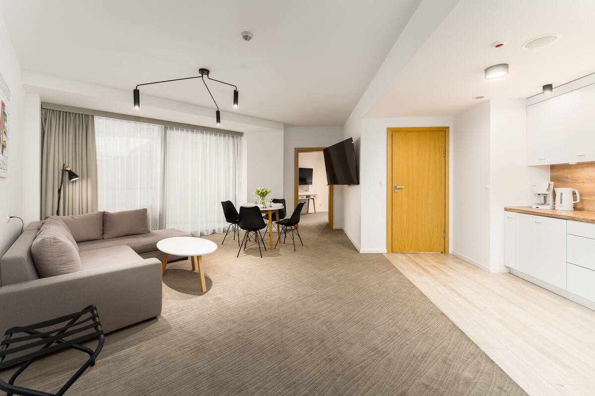 A modern living room with a sofa, dining table, and chairs. There is a kitchen area with cabinets and a countertop.