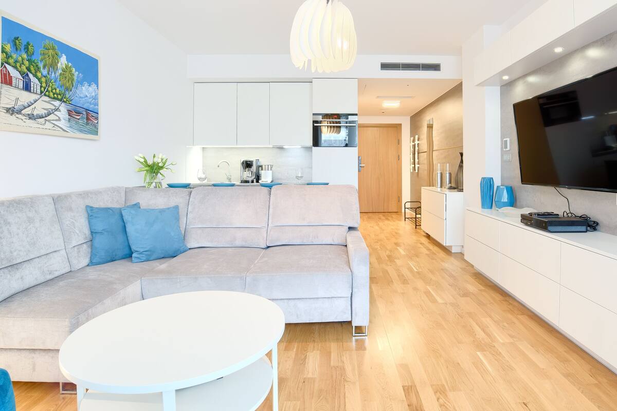 A modern living room with a grey sofa, a round white table, and a kitchen area with white cabinets and a flat-screen TV.