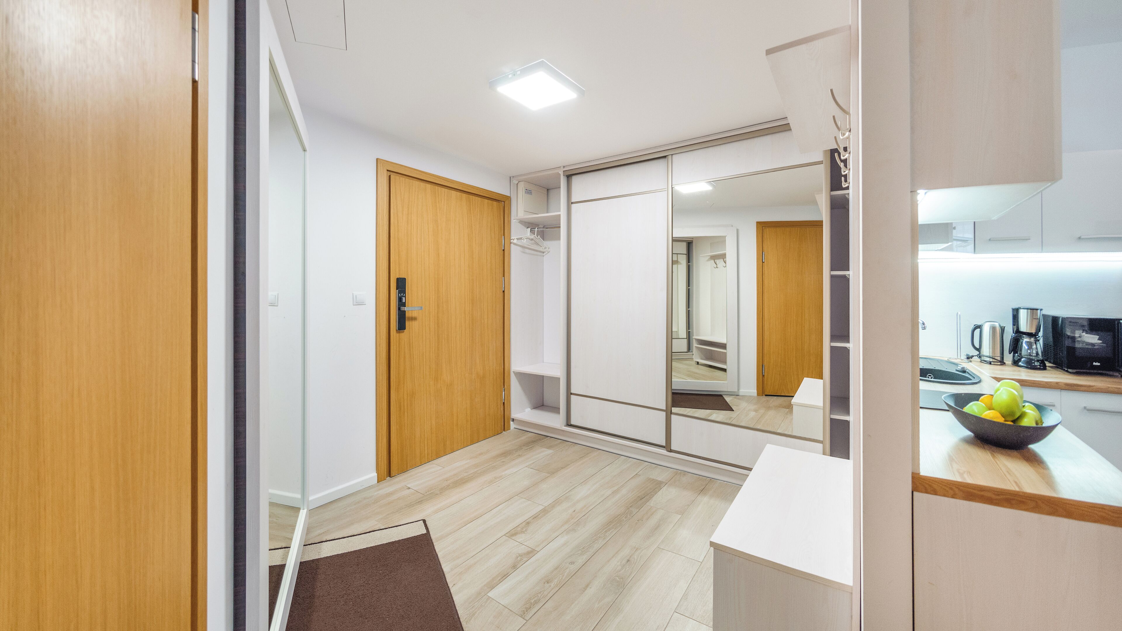 Une chambre d’hôtel moderne avec une porte en bois, une armoire, un miroir, une kitchenette équipée d’une machine à café et un panier de fruits posé sur le comptoir.