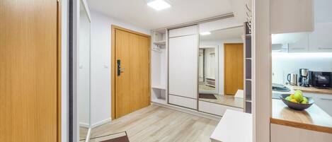 Une chambre d’hôtel moderne avec une porte en bois, une armoire, un miroir, une kitchenette équipée d’une machine à café et un panier de fruits posé sur le comptoir.