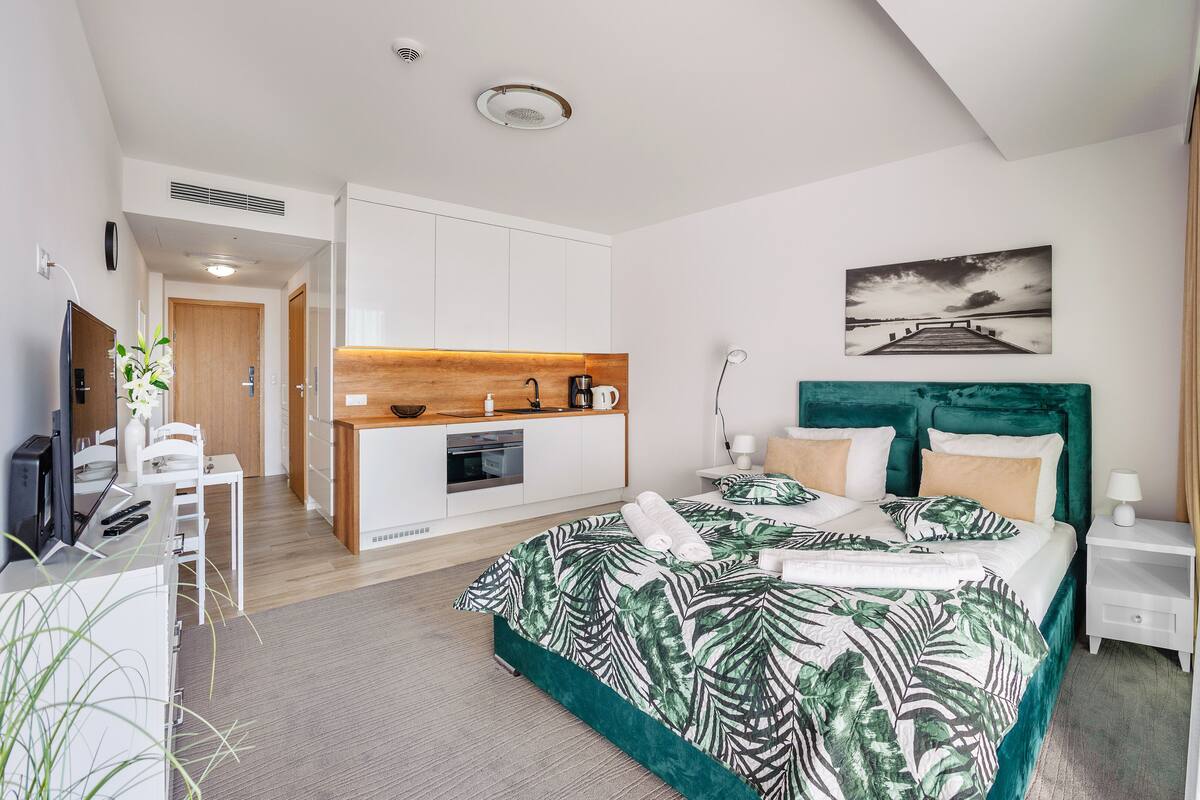 A modern hotel room with a large bed, a kitchenette, a desk, and a view of the sea.