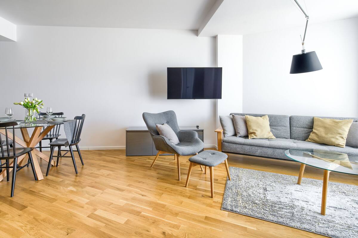 A modern living room with a dining area, a grey sofa, a flat-screen TV, and a wooden floor.