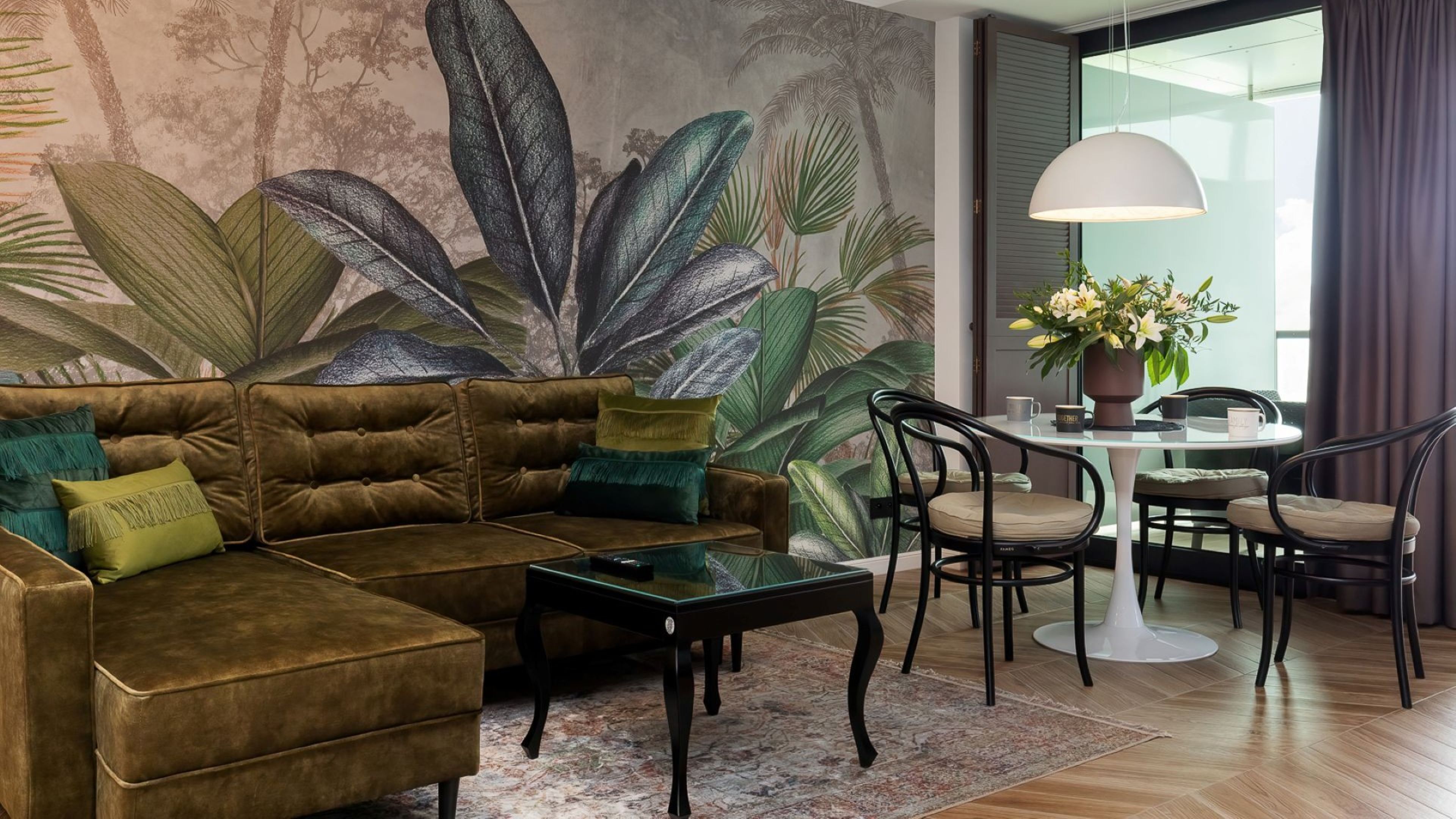 Una sala de estar con un sofá marrón, cojines verdes, una mesa de centro de vidrio y un mural de hojas tropicales. Incluye una mesa de comedor con sillas y una planta en maceta.