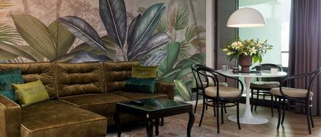 Una sala de estar con un sofá marrón, cojines verdes, una mesa de centro de vidrio y un mural de hojas tropicales. Incluye una mesa de comedor con sillas y una planta en maceta.