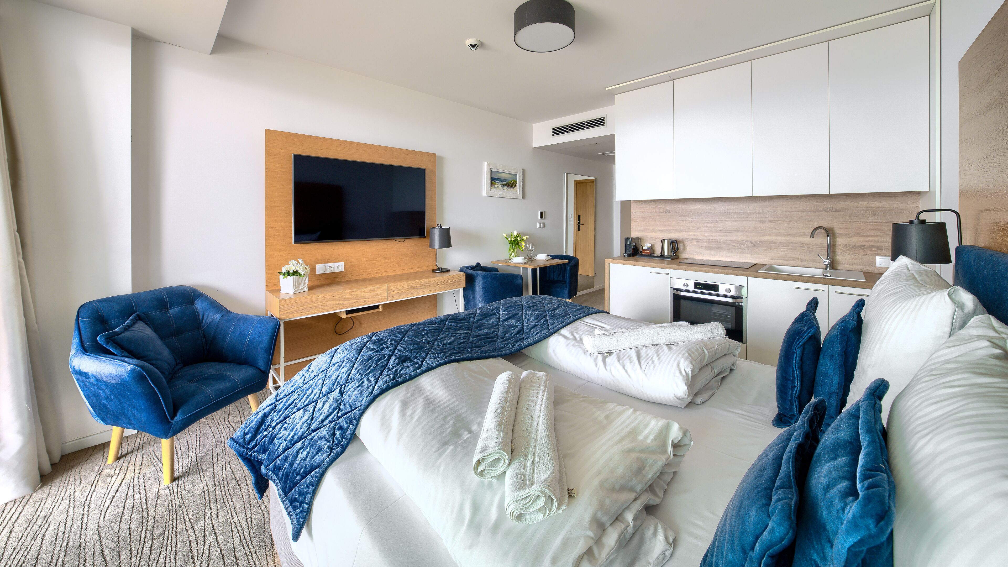 A modern hotel room with a bed, a desk, a chair, a TV, a kitchen area, and a bathroom.
