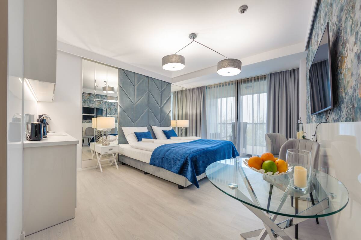A modern bedroom with a bed, bedside tables, a glass table with fruits, and a TV on the wall.