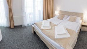 A neatly made bed with white linens and beige pillows, a bedside table with a lamp, and a view through a window with sheer curtains. - Hotel Prístav (Trstená)