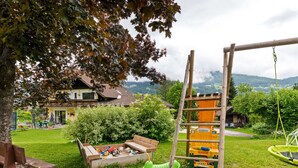 Ein Spielplatz mit Schaukeln und einer Rutsche, umgeben von einer Rasenfläche mit einer Bank und einem hölzernen Spielhäuschen.