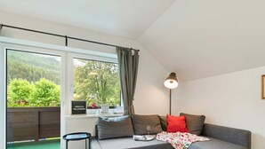 Ein modernes Wohnzimmer mit einer grauen Eckcouch, einem kleinen Beistelltisch, einer Stehlampe und einem großen Fenster mit Blick ins Grüne.