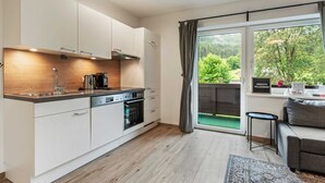 Eine moderne Küche mit weißen Schränken, einem eingebauten Ofen und einer Kaffeemaschine. Ein Essbereich mit einer grauen Couch und einem kleinen Tisch. Ein großes Fenster bietet Blick auf Grünflächen.
