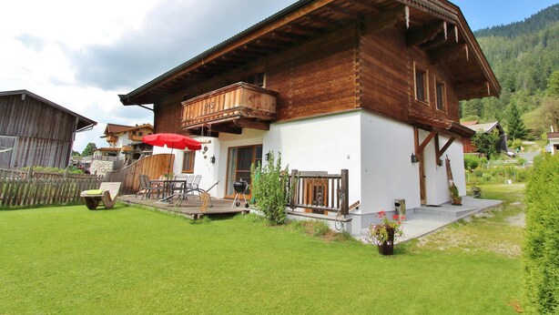 A two-story wooden house with a balcony, a red umbrella, and a well-maintained lawn.
