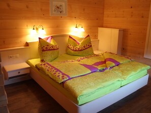Uma cama com colchão verde, duas almofadas e um cobertor estampado. Há duas mesas de cabeceira com luminárias e uma pequena gaveta.