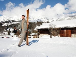 Eine Person trägt Ski und einen Helm und geht auf einem verschneiten Pfad. Im Hintergrund sind eine Holzhütte und Berge zu sehen.