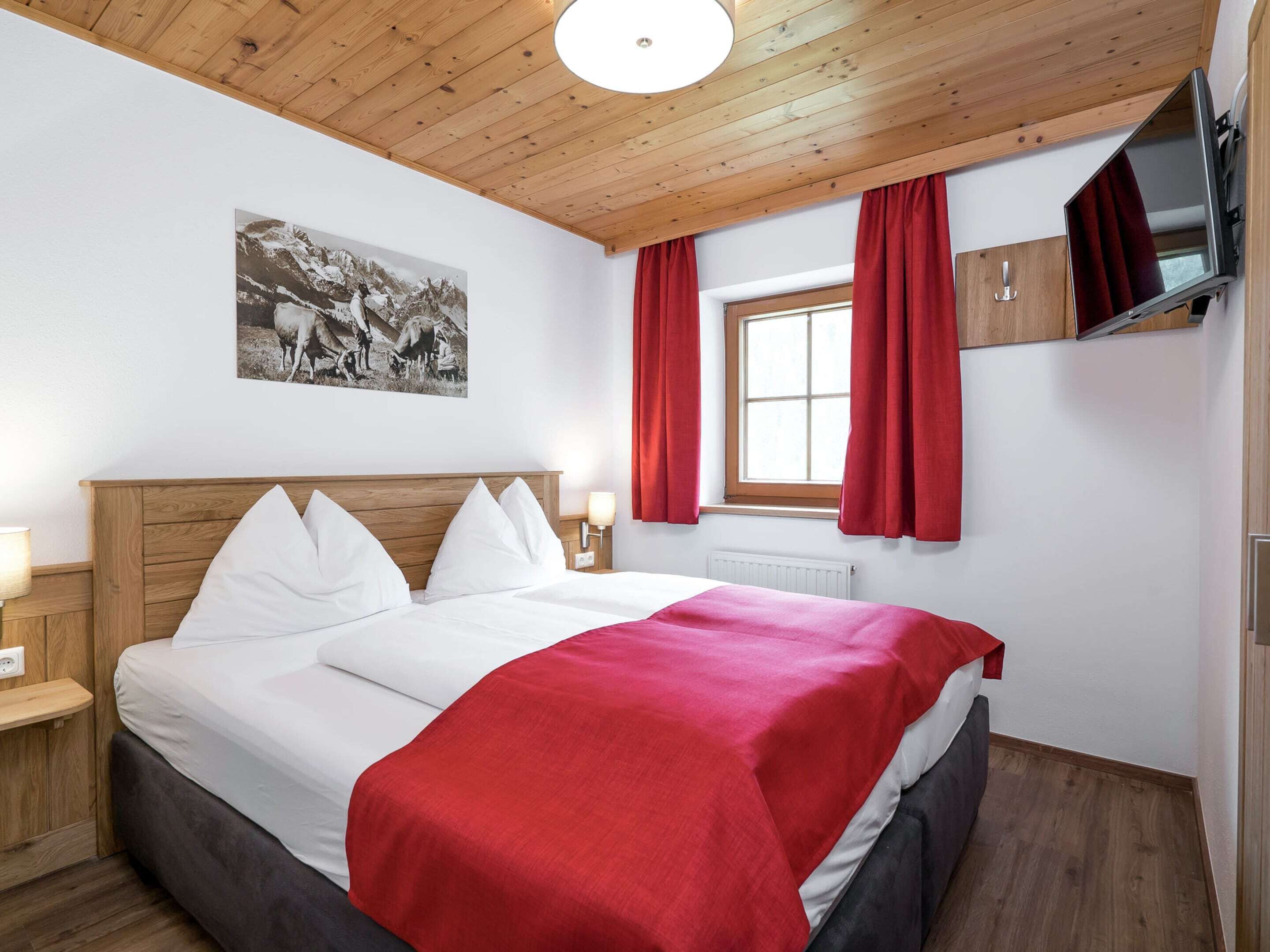 Une chambre d’hôtel avec un plafond en bois, un lit avec une literie blanche et rouge, un téléviseur fixé au mur et une fenêtre avec des rideaux rouges.