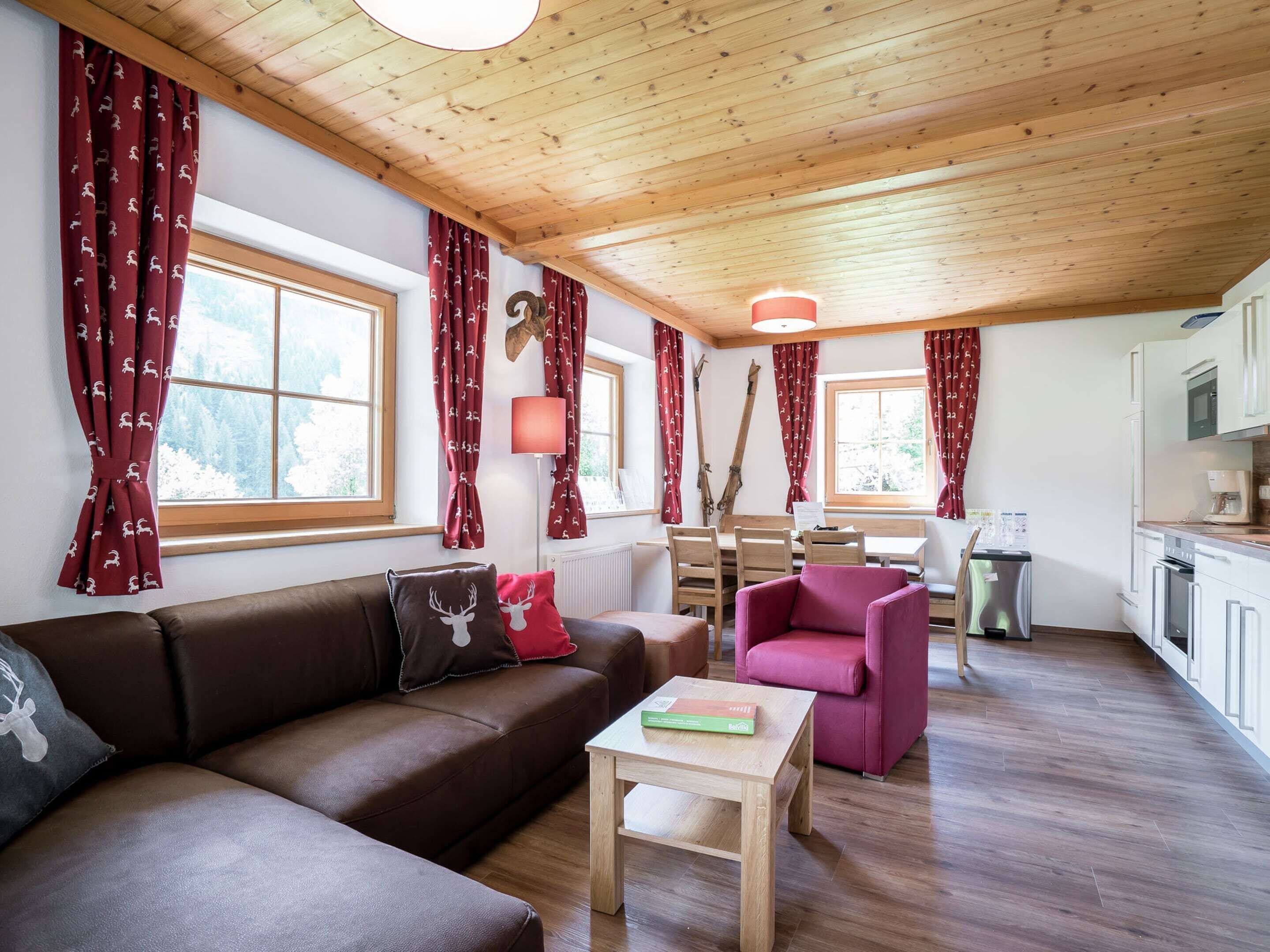 Un salon chaleureux avec un canapé marron, un fauteuil rose et une table basse en bois. On y trouve également un coin repas avec une table et des chaises en bois, ainsi qu’une cuisine dotée de meubles et d’appareils électroménagers blancs.