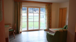 A room with a green sofa, a wooden table, and a chair. There is a large window with curtains and a view of a grassy area.