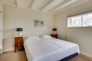 Una cama bien hecha en una habitación con suelo de madera, dos mesitas de noche con lámparas, una ventana con persianas y un cuadro colgado en la pared.