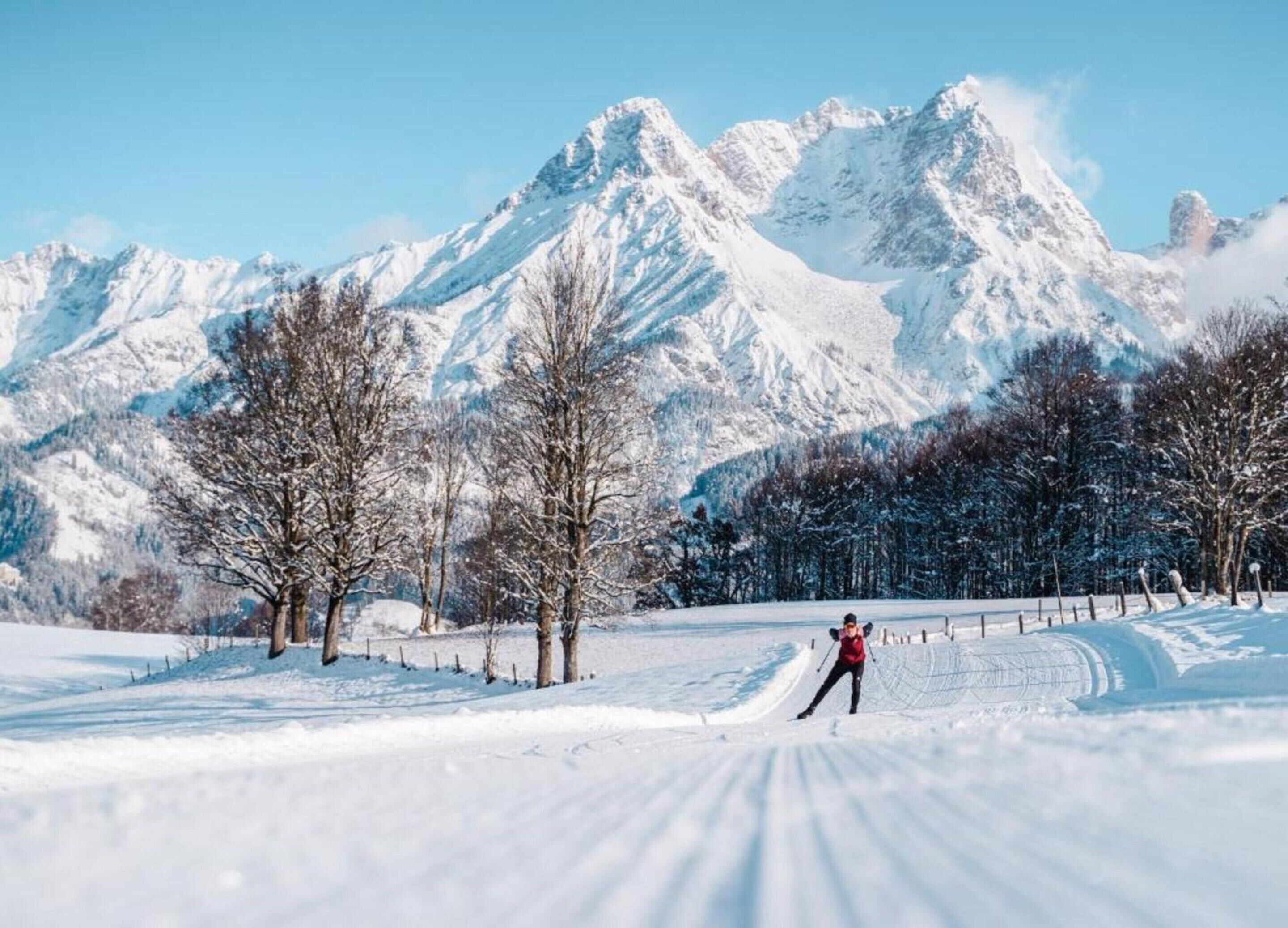 A snowy landscape with a person cross-country skiing, trees, and snow-covered mountains in the background.