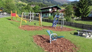 A playground with swings and slides, surrounded by a fenced area with a bench and mulch. - Apartment With Mountain View (Piesendorf)
