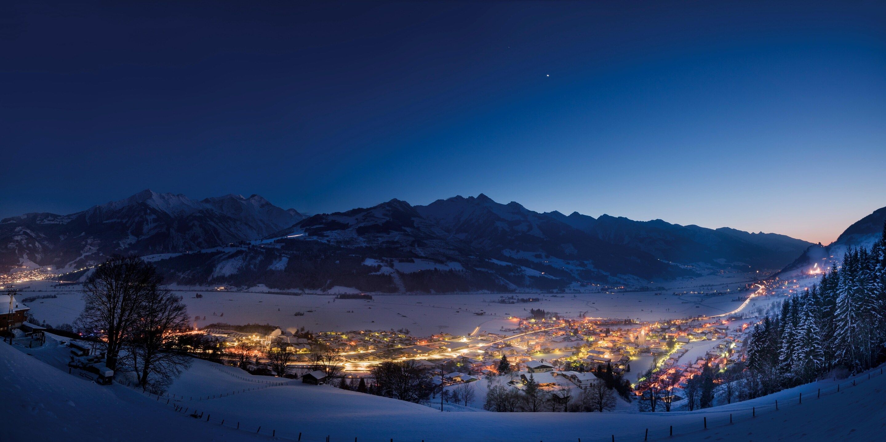A snow-covered mountainous landscape at twilight with a lit town in the valley.