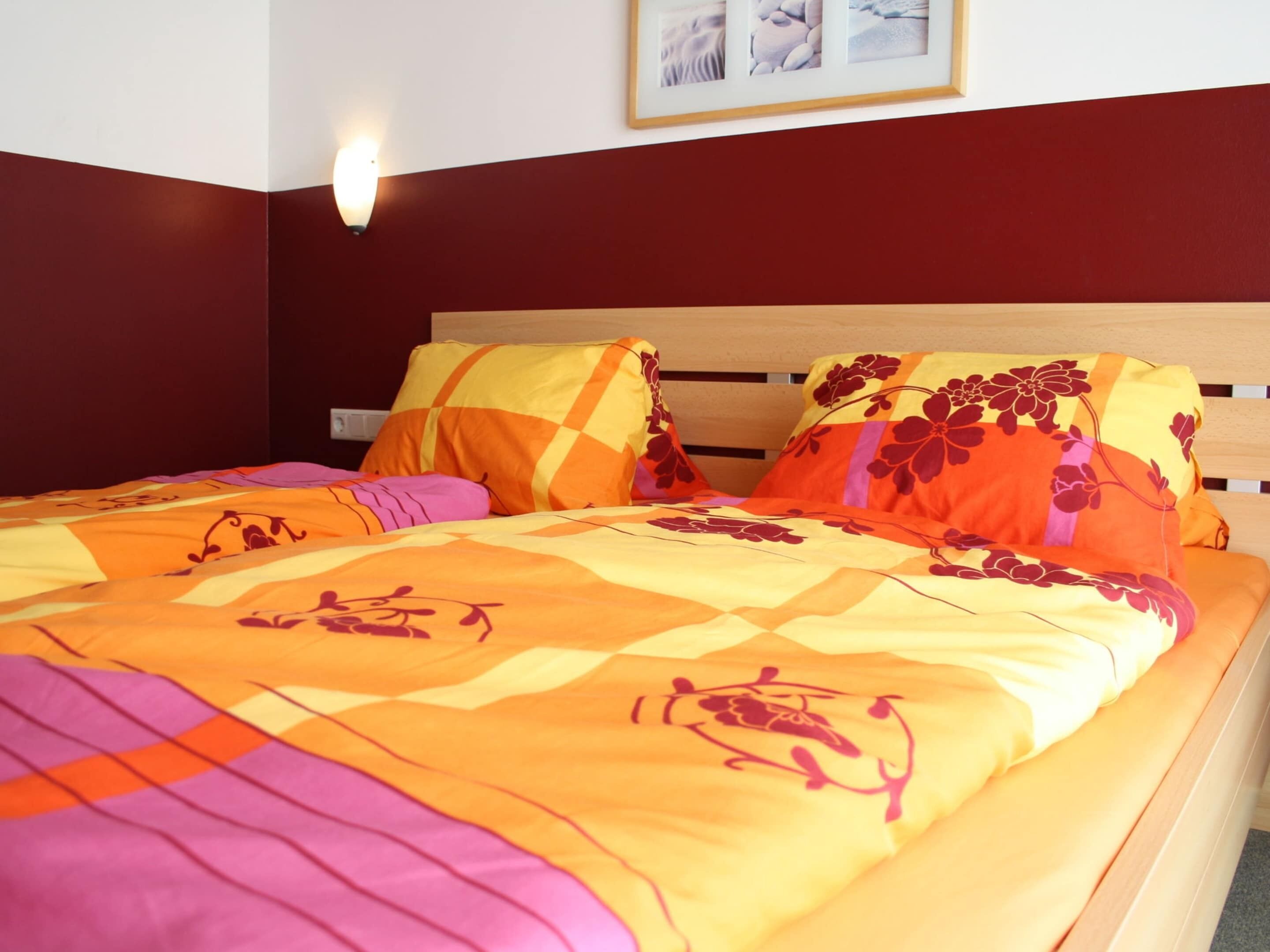 A bed with a floral-patterned bedspread and matching pillows, a wooden headboard, and a wall-mounted light fixture.